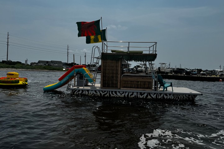 Boat with slide and flags on water, inflatable nearby, overcast sky.